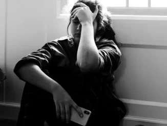 En tonåring sitter med ryggen mot en vägg ledsen med en telefon i handen.