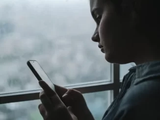 tonåring står vid ett fönster och tittar i mobilen,